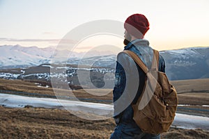 Bearded tourist hipster man in a hat with a backpack stand back on a roadside bump and watching the sunset against the