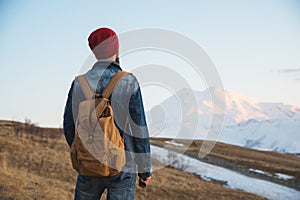 Bearded tourist hipster man in a hat with a backpack stand back on a roadside bump and watching the sunset against the