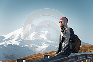 Bearded tourist hipster man in sunglasses with a backpack sitting on a roadside bump and watching the sunset against the