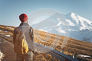 Bearded tourist hipster man in sunglasses with a backpack stand back on a roadside bump and watching the sunset against