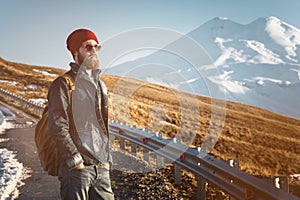 Bearded tourist hipster man in sunglasses with a backpack stand back on a roadside bump and watching the sunset against