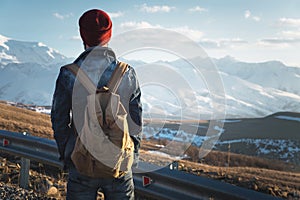 Bearded tourist hipster man in sunglasses with a backpack stand back on a roadside bump and watching the sunset against