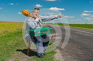 bearded Ukrainian senior gestures while sitting on summer roadside with an ancient green suitcase and mandoli