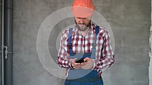 bearded worker or builder in safety helmet using smartphone over grey wall background. Profession, construction and