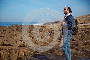 Bearded young adult man contemplating the nature and seascape, standing on the cliff, carrying his travel backpack