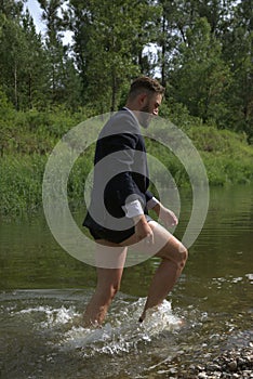 Bearded young businessman in black classic suit jacket, boxer underwear run ankle-deep in water of narrow forest river