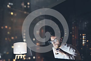 Bearded young businessman working on modern loft office at night. Man using contemporary notebook texting message, holding cup esp