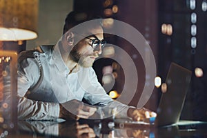 Bearded young businessman working on modern loft office at night. Man using contemporary notebook texting message, holding smartph