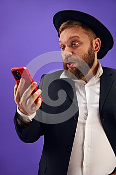 Bearded young Jewish man emotionally looking on mobile phone screen, reading news and messages against purple studio
