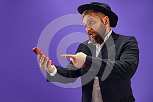 Bearded young Jewish man emotionally looking on mobile phone screen, reading news and messages against purple studio