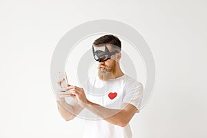 Bearded young male wears batman mask, uses smart phone for making photo, poses against white blank studio wall. Handsome