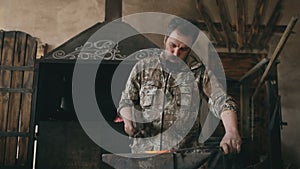 Bearded young man blacksmith manually forging hot metal on the anvil in smithy with spark fireworks