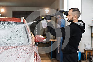 Bearded young man car wash worker spraying cleaning foam to a modern red car