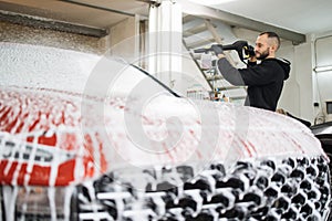 Bearded young man car wash worker spraying cleaning foam to a modern red car