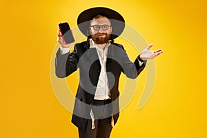 Bearded young man in hat with sidelock holding mobile phone, greeting with holiday against yellow studio background