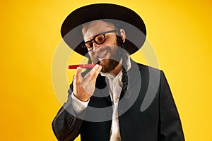 Bearded young man in hat with sidelock recording voice message on phone, greeting with holiday against yellow studio