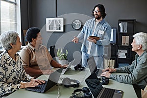 Bearded young man leading computer class for seniors using laptops at table