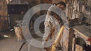 Bearded young man using angle grinder to work on piece of metal in smithy workshop, grinding sparks flying around