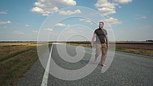 Bearded Young Man Wearing Glasses, T-shirt and Shorts Walking Barefoot on a Deserted Automobile Road Between Fields on a