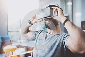 Bearded young man wearing virtual reality goggles in modern coworking studio. Smartphone using with VR headset in office