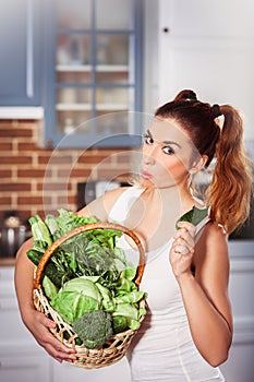 Beatiful caucasian girl in sporty wear holding basket of fresh vegetables and chews cucumber.