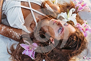 Beatiful young woman lying on sand with pink and white lilies on the beach at sunset