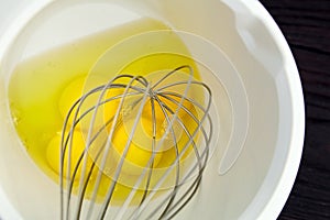 Beating raw yellow egg yolks with a handle steel whisk in white bowl on wooden table. Mixing food ingredients. Cooking chicken
