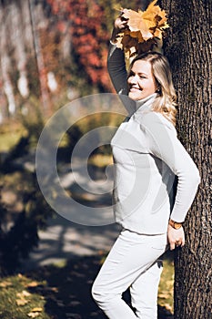 Beauteous smiling middle-aged woman with long wavy fair hair lifting hand with bouquet of fallen leaves leaning on tree.