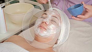 Beautician applies a massage cream on the face of an Asian middle-aged woman. Rejuvenating procedure in the beauty salon. White co