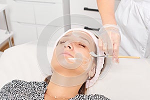 Beautician applying cosmetic cream mask on woman face for rejuvenation, procedure in beauty salon