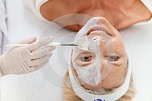 Beautician applying cosmetic mask on half woman face with cosmetic brush at beauty salon