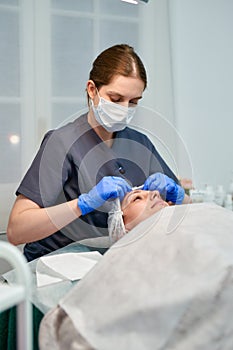 Beautician is applying healthy cream on female facial skin. Woman is lying on table