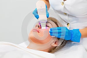 A beautician in blue medical gloves does an ultrasound facial cleansing for an adult Caucasian woman. Cleansing