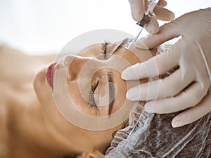 Beautician doing beauty procedure with syringe to face of young brunette woman in sunny clinic. Cosmetic medicine and