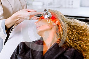 Beautician doing red led light therapy to blond woman in beauty salon, facial photo therapy for skin pore cleansing