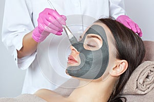 Beautician makes a black clay mask to rejuvenate the skin to beautiful woman.Cosmetology concept and treatment of problem skin