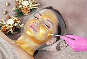 Beautician makes a golden mask to rejuvenate the skin to beautiful woman. Next to her are Christmas decorations.New Year`s and