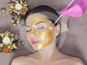 Beautician makes a golden mask to rejuvenate the skin to beautiful woman. Next to her are Christmas decorations.New Year`s and