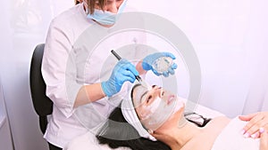 Beautician in a medical mask and gloves with a silicon brush applies a white moisturizing facial mask to the face of a