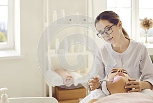 Beautician at spa salon applying mask with pink clay on face of relaxed young woman