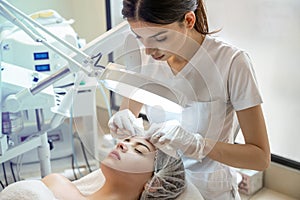 Beautician squeezing acne and pimples from young woman& x27;s nose using lamp and magnifying glass in beauty spa clinic.