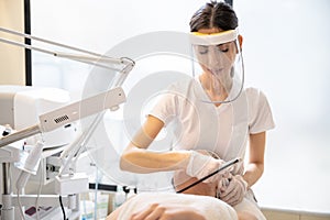 Beautician wearing Face shield doing rejuvenation treatment on woman face in beauty salon.