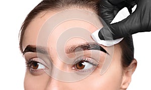 Beautician wiping tint from woman`s eyebrows on white background, closeup photo