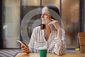 Beautifil charming happy businesswoman in formal attire while using smartphones drinking coffee in cafe sitting with mobile phone