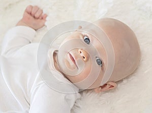 Beautiful 6 Month Baby Boy Dressed in White & Lying on Fluffy White Blanket Looking at Camera. Smiling & Happy