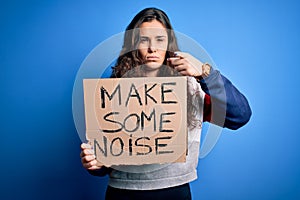 Beautiful activist woman holding banner with make some noise message over blue background pointing with finger to the camera and