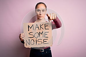 Beautiful activist woman holding banner with make some noise message over pink background pointing with finger to the camera and