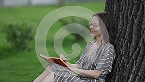 Beautiful adult student doing her college homework sitting on the grass in the city park. A woman learns outdoors