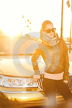 Beautiful adult woman in leather jacket and scarf in golden sunlight near car