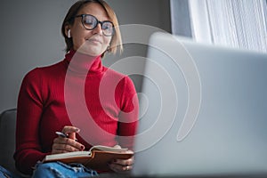 Beautiful adult woman psychologist wearing glasses at home sitting at home on the couch in front of laptop screen taking notes